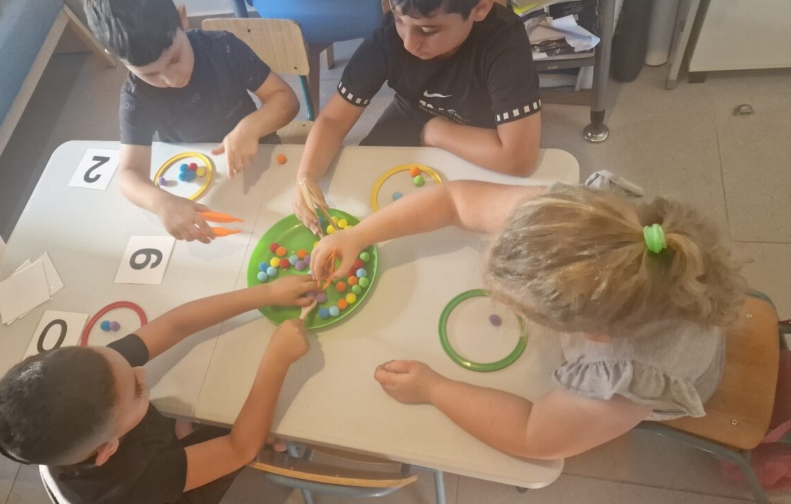 ילדים מתרגלים מספרים וצבעים סביב שולחן בקבוצת הכנה לכיתה א׳ במרכז רגב